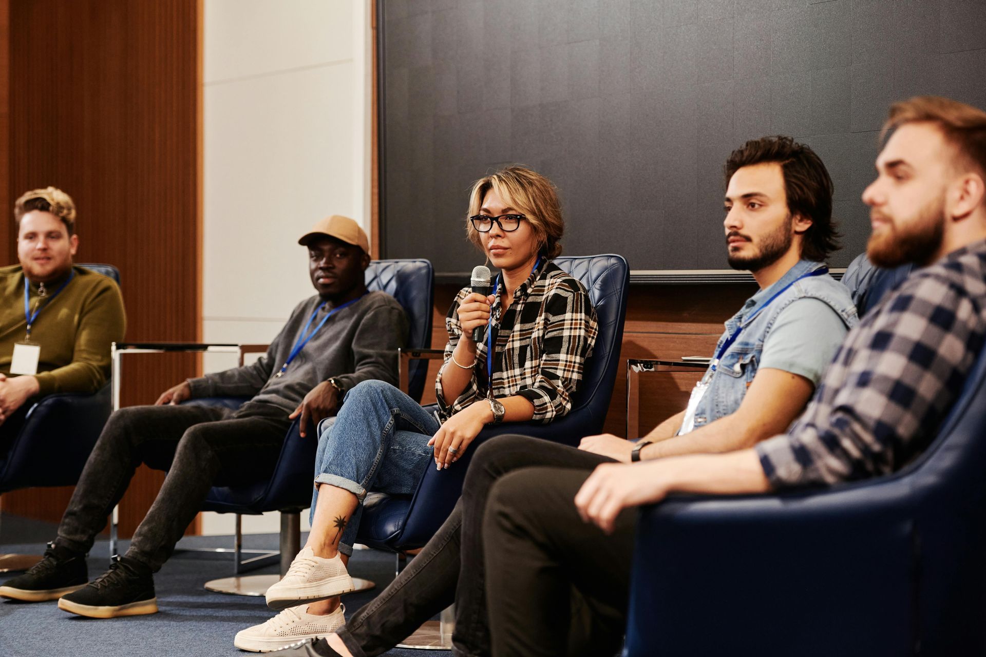 Five people sit in blue chairs on a stage, with one person speaking into a microphone during a professional panel.