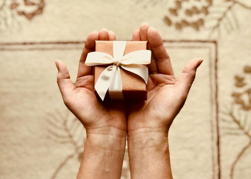 Two cupped hands holding a small, brown square gift box wrapped with a white ribbon against a patterned fabric background.