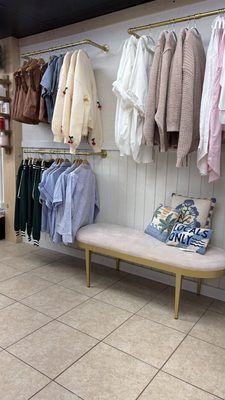 Clothing racks display sweaters and shirts in a boutique, with a cushioned bench and throw pillows underneath.