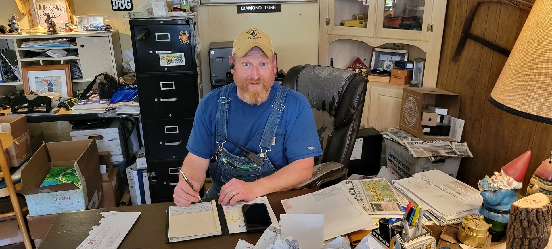 A man in overalls sits at a desk in a cluttered room