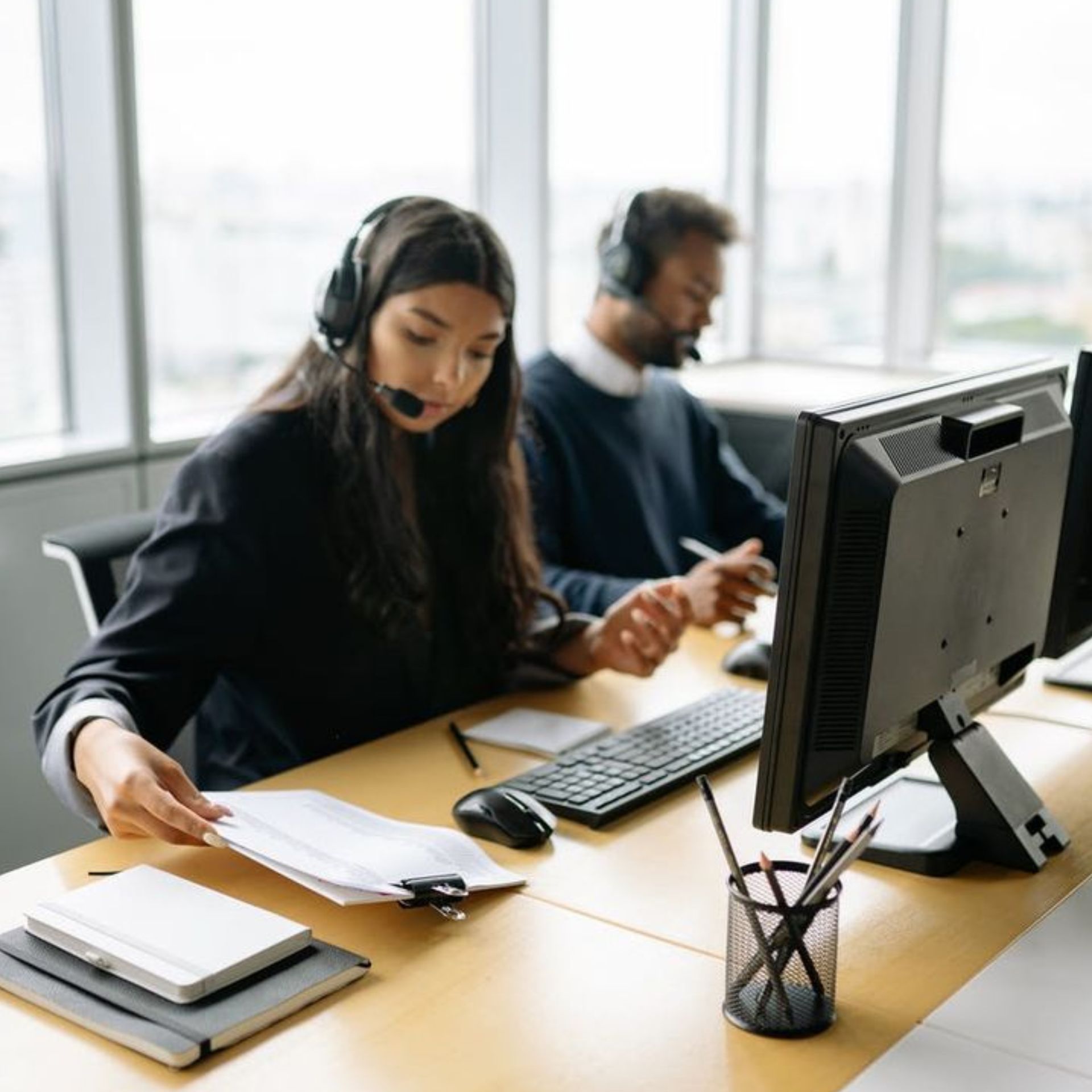 Two office workers wearing headsets at desks with computers in a bright workspace.