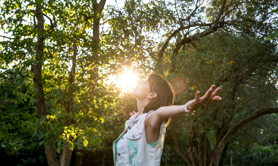 Woman writing in a notebook outside, smiling at the page. Golden sunlight, pink cardigan.