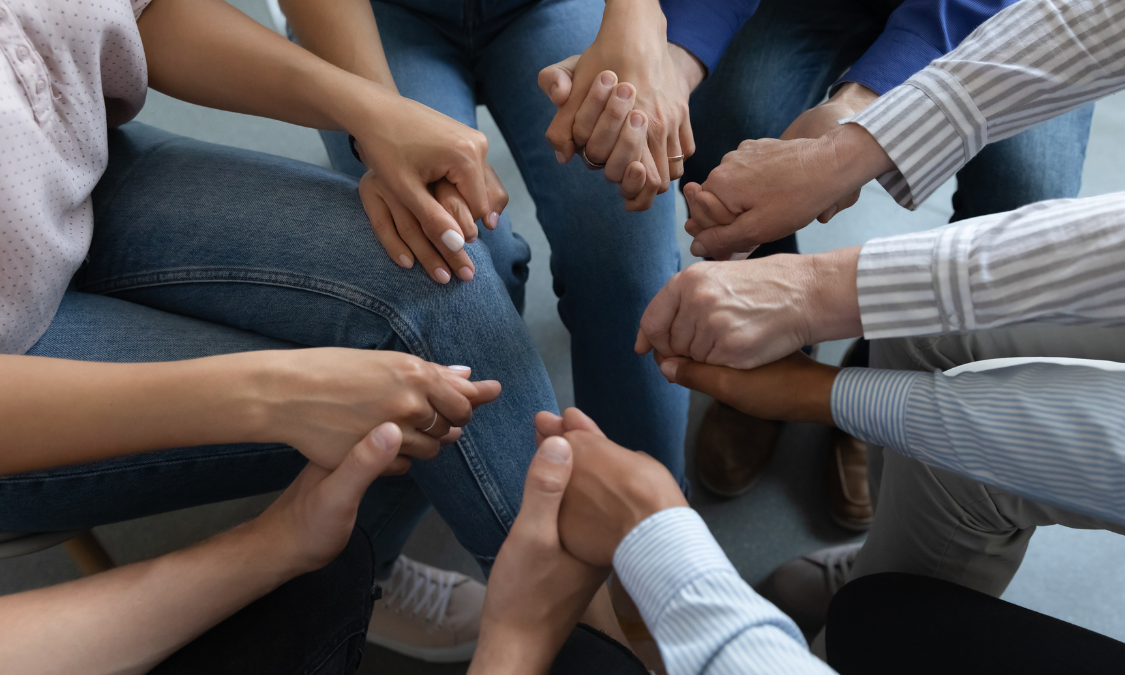 Person's clasped hands in foreground, blurred figures in background, inside setting, possibly a therapy session.