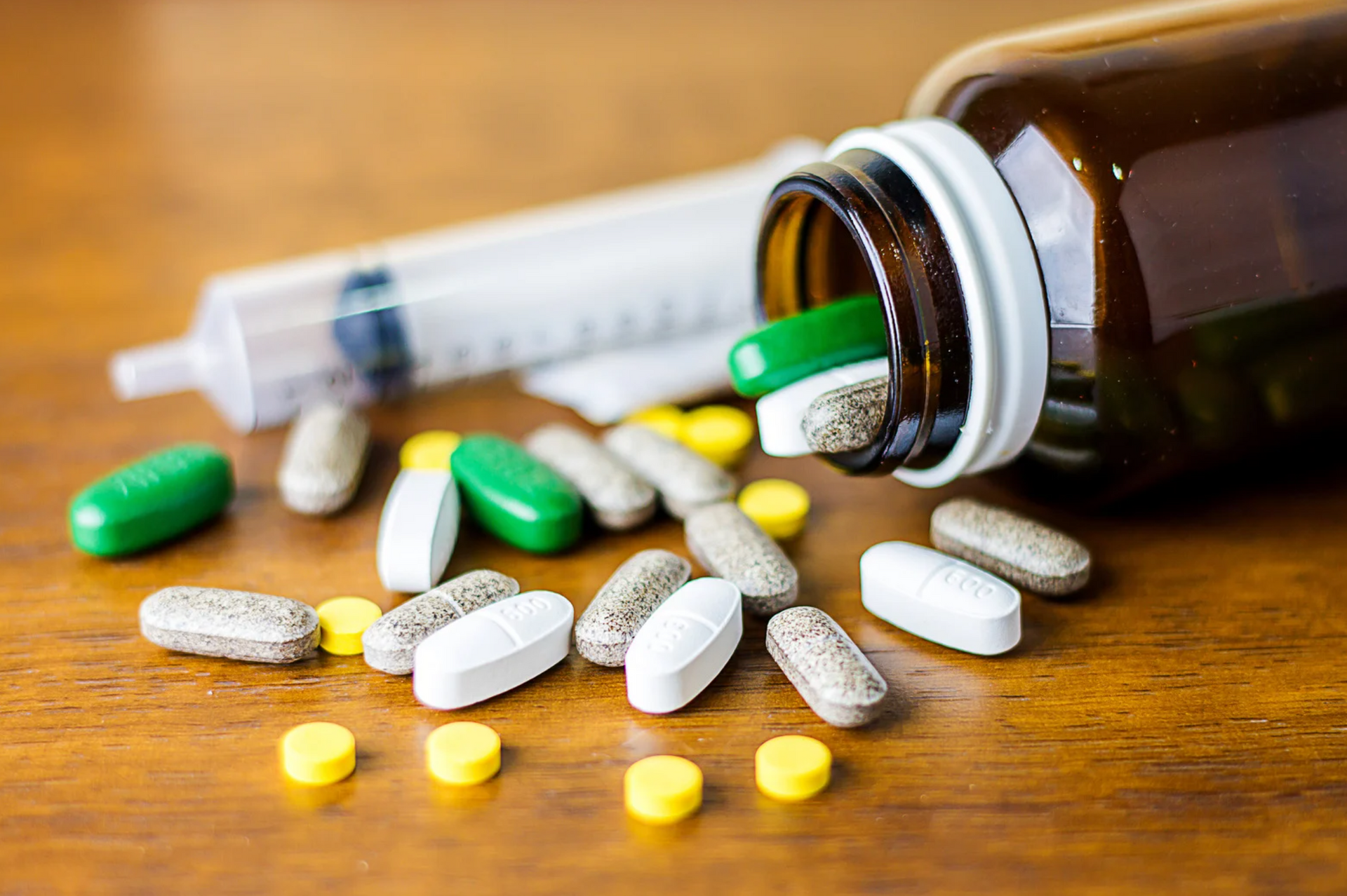 Pills spilling from a bottle with a syringe on a wooden surface.
