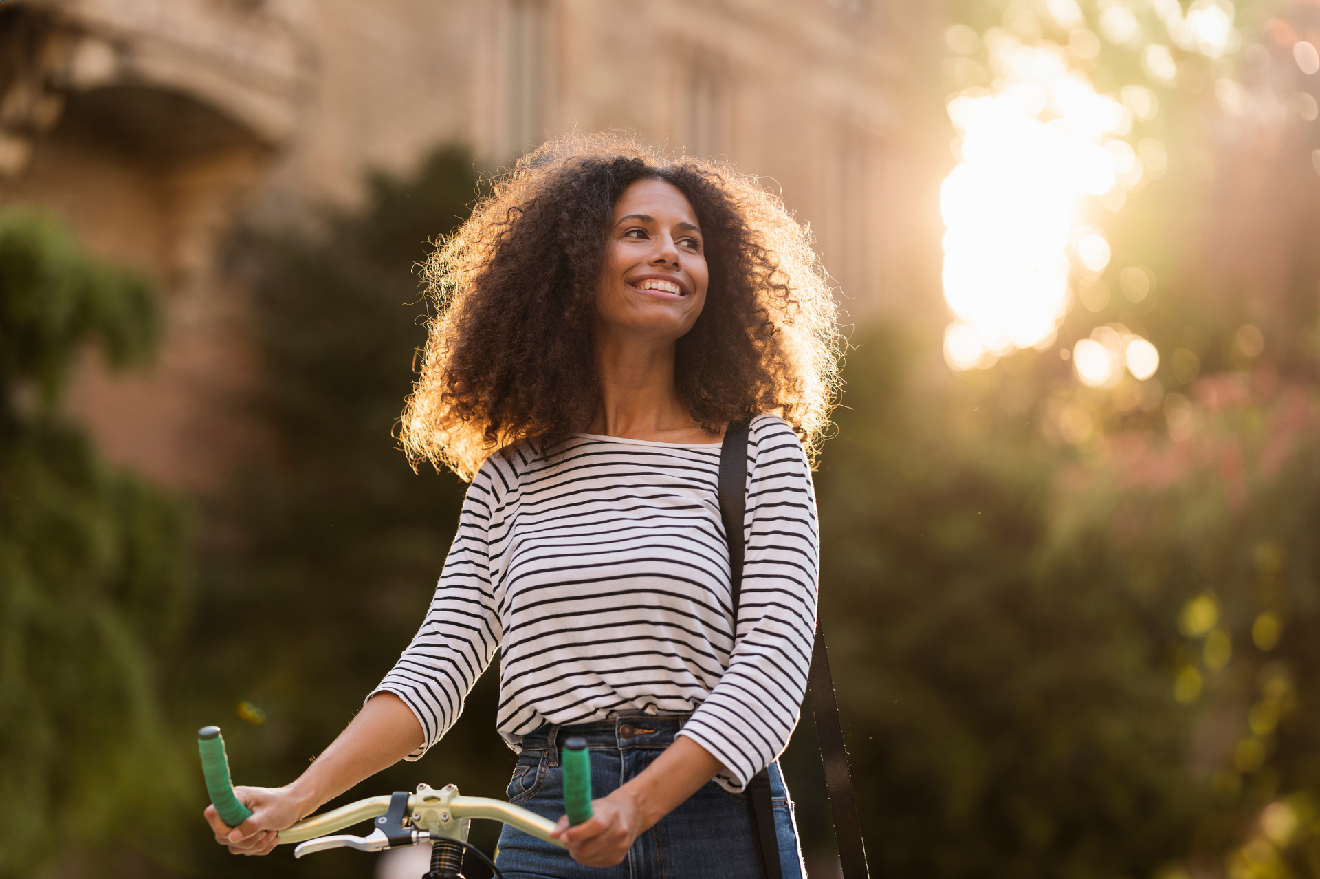 Woman with curly hair smiles, holding bike, outdoors in the sun.