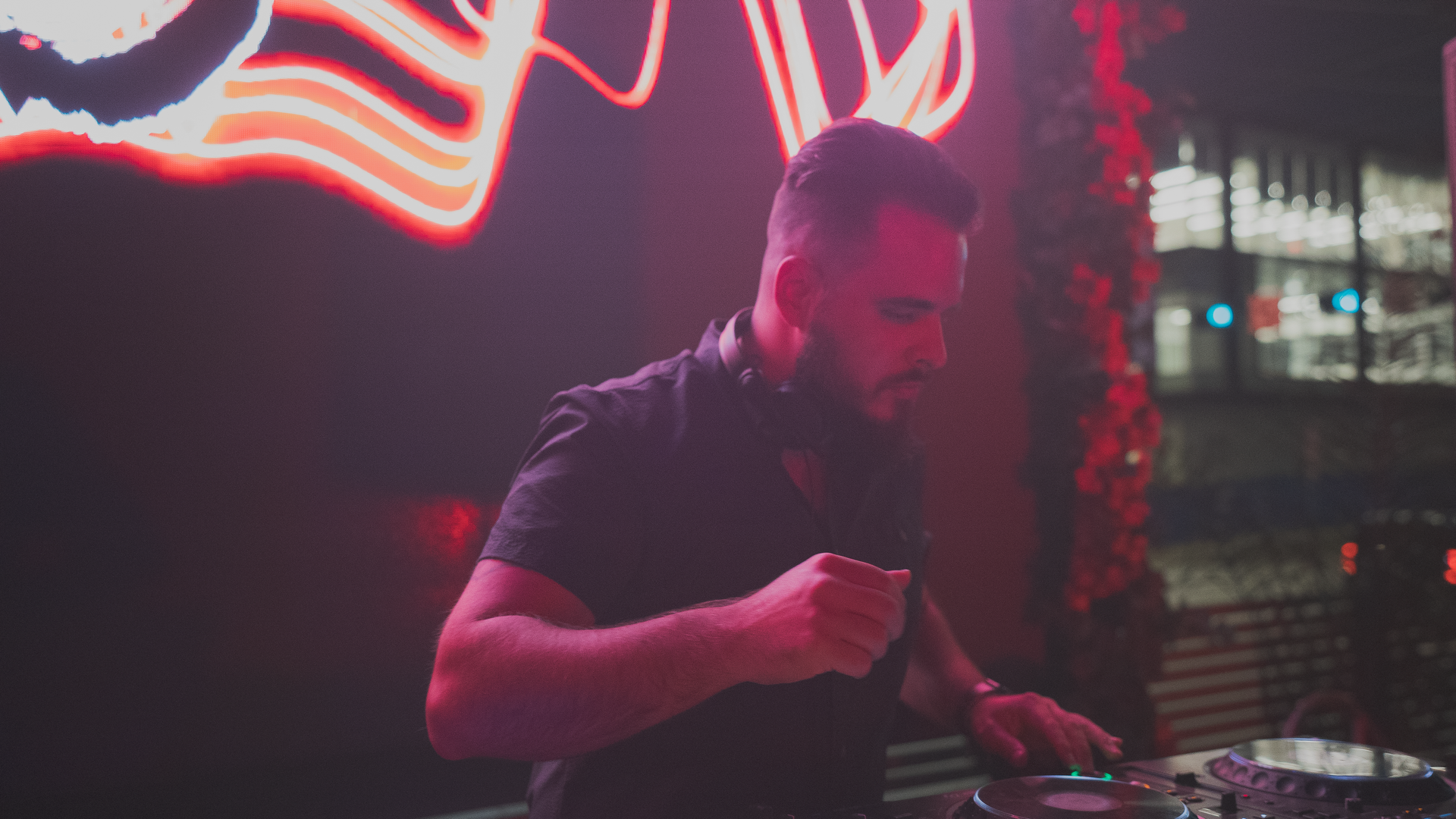 DJ wearing headphones, performing at an event. Laptop and mixing console are visible. Red lighting in the background.