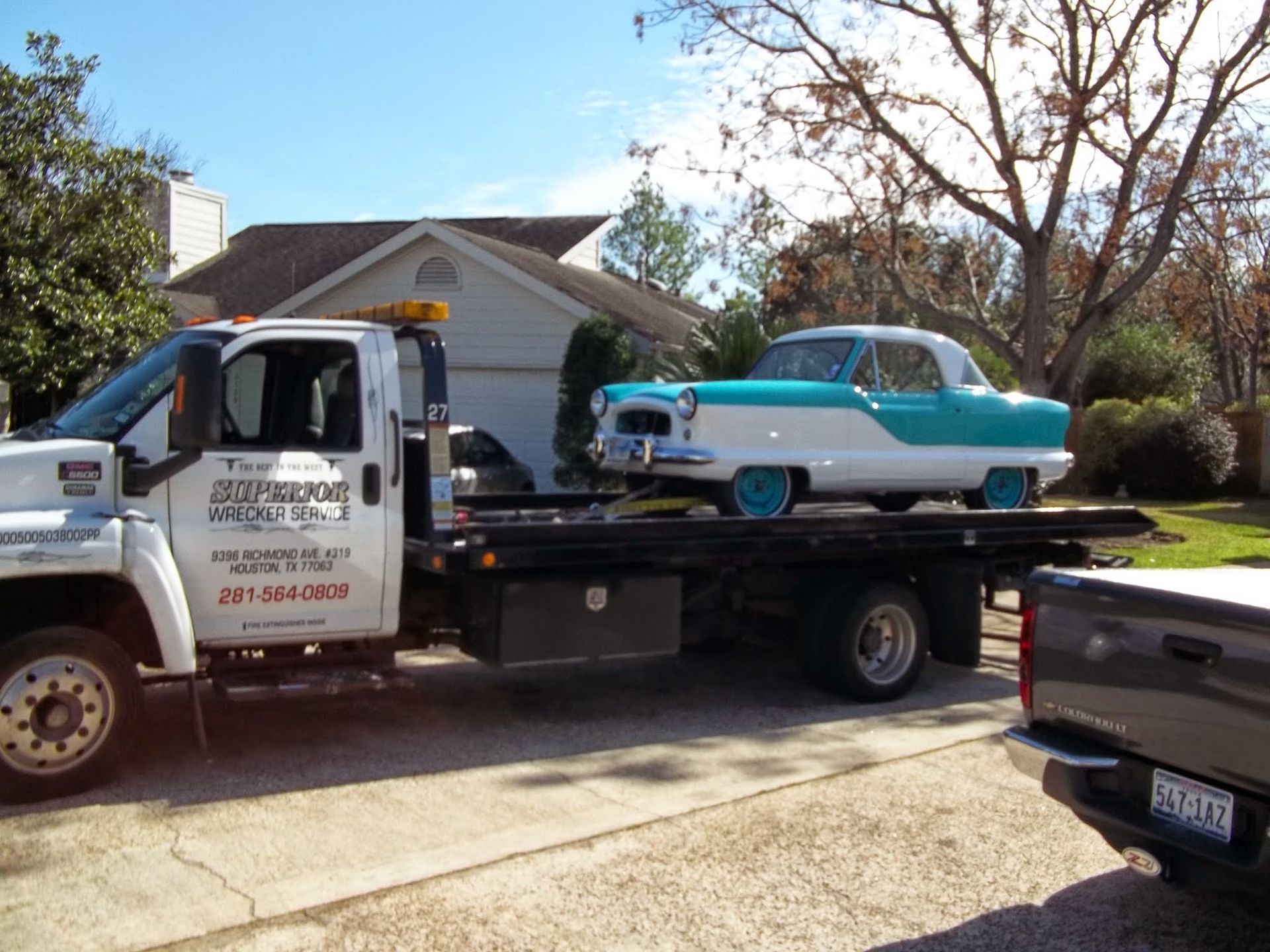 A vintage teal and white car on a tow truck in front of a house
