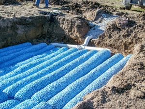 A septic drain field under construction with blue synthetic aggregate modules and white PVC piping in an excavated trench.