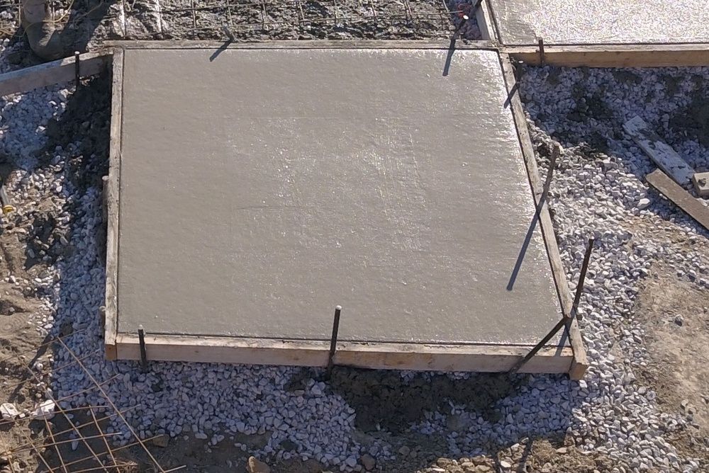A freshly poured concrete slab in a wooden frame, surrounded by gravel and soil at a construction site.