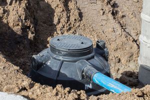 A black plastic septic or distribution basin with a blue pipe attached, partially buried in a dirt trench.