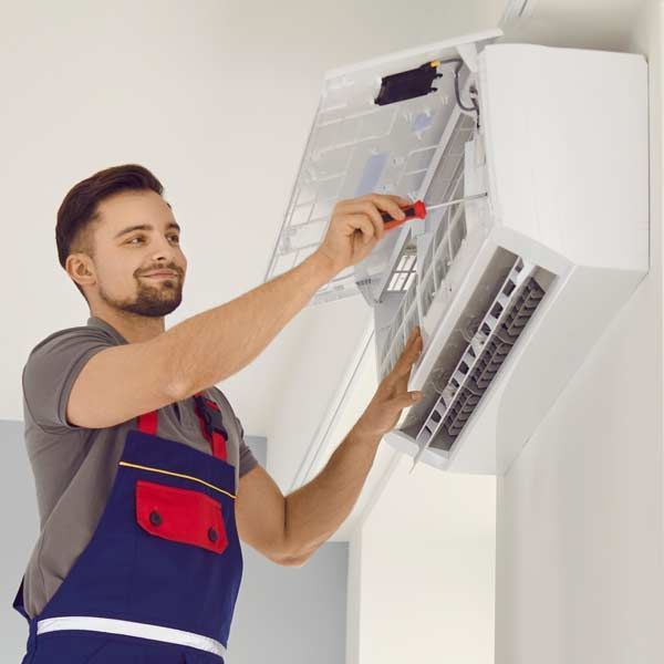 A man in overalls smiles, repairing a white air conditioner unit with a screwdriver. Indoor, white wall representing Air Conditioner Repairs and Maintenance