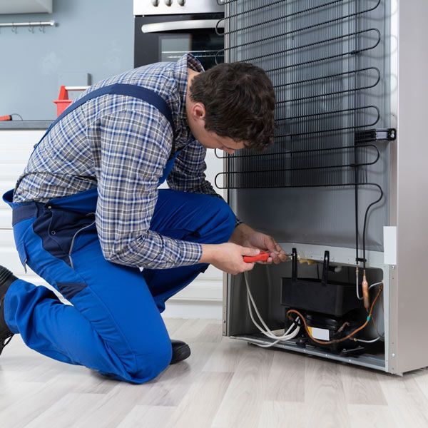 A man in blue coveralls is repairing the refrigerator's compressor indoors. He is using a screwdriver on his Appliance Repair Services