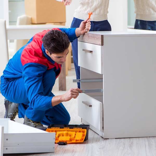 A man in a blue jumpsuit assembles a white dresser with a toolbox on the floor. Woman in background watching the Cabinet Repair Services