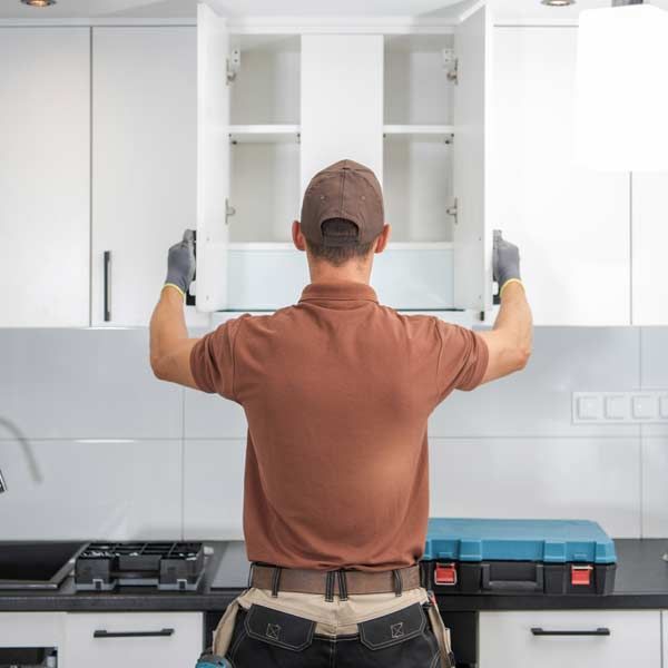 A man is installing a kitchen cabinet, wearing gloves, facing the cabinets, with a brown shirt and a toolbox nearby.