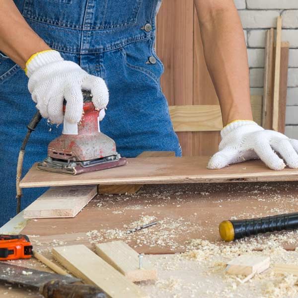 Carpenter sanding a wooden board with an electric sander, wearing gloves and overalls in a workshop, providing Carpentry Services