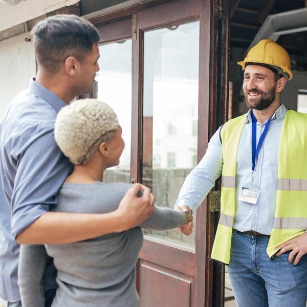 Couple greets contractor at a doorway. The worker shakes hands and wears a hard hat and safety vest.
