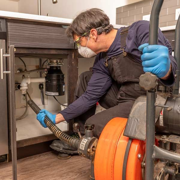 A handyman is using a drain snake under a kitchen sink. Wearing gloves, mask, and safety glasses for the Clogged Drain Repair Services