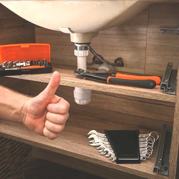 Albert Sindoni, the Honest Hammer Handyman in Oceanside, Nassau County, Long Island, gives a thumbs up under a bathroom sink with tools, a cabinet, and plumbing after repairing a clogged drain