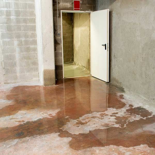 A clogged floor drain causes a flooded basement with an open white door, a wet red floor, and concrete walls.