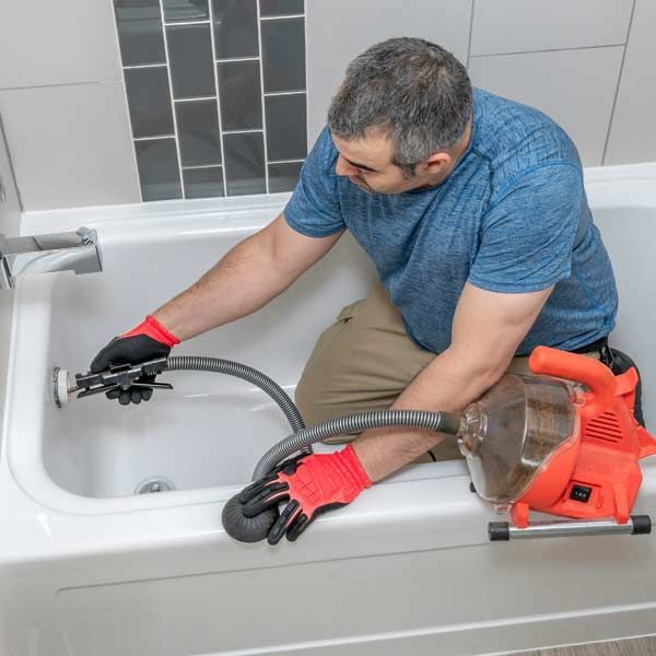 A handyman is repairing a clogged shower and bathtub using a drain snake in a white bathtub, wearing gloves, in a bathroom.