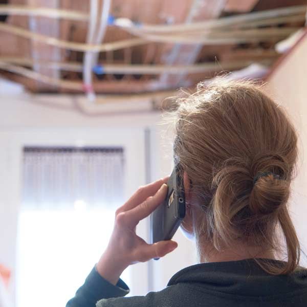 A woman on the phone looks at the water-damaged ceiling, with visible damaged beams and wires, possibly requesting repairs indoors Contacting Albert Sindoni for a Free Renovation Consultation