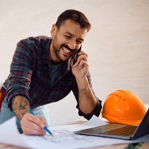 Construction worker, smiling, on phone, drawing plans, orange hard hat, laptop on desk. Contacting Albert Sindoni of Honest Hammer for a Free Consultation on Subcontracting Services in Nassau and Suffolk County