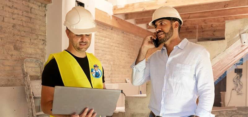 Two construction workers in hard hats, one on a phone, the other looking at a laptop in a building under construction. Contractor Subcontractor Services
by Honest Hammer