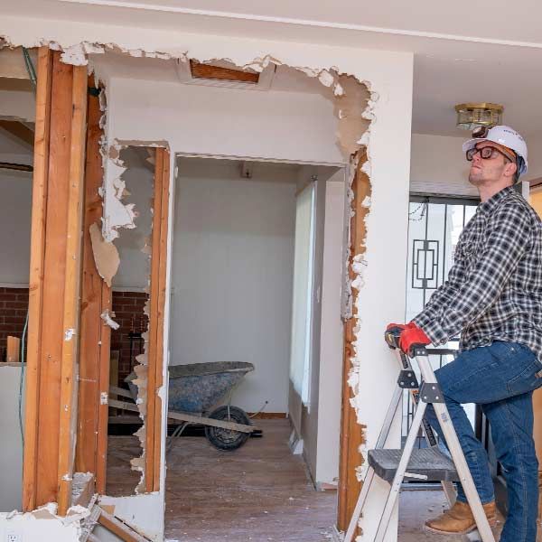 Man in hard hat inspects a wall opening during a home renovation Demolition; ladder, debris, and wheelbarrow are visible.