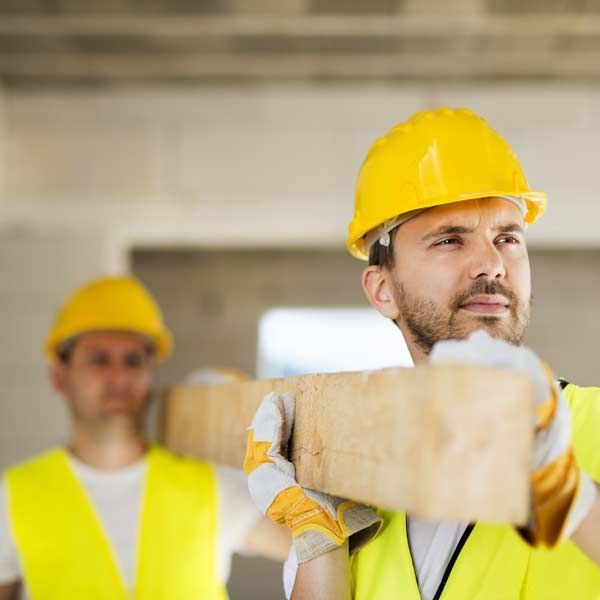 Two construction workers wearing yellow vests and hard hats carrying wooden planks.