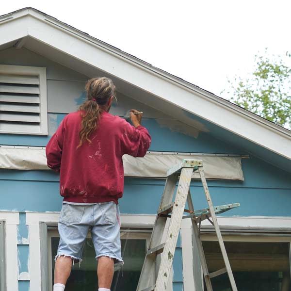 Contractor Exterior Painting blue house trim while standing on a ladder.