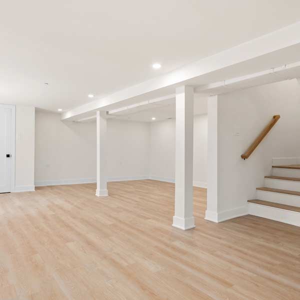 Empty, bright finished basement with wood floors, support beams, and a staircase.
