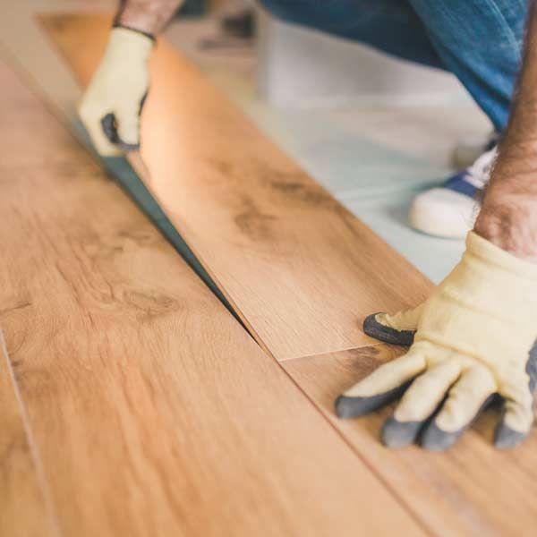 Person kneeling, installing wooden floorboards, wearing gloves and jeans doing Flooring Installation and Repair