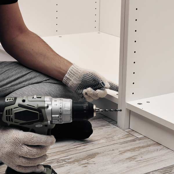 A person using a power drill to assemble a white cabinet, wearing work gloves while doig Furniture Assembly Services