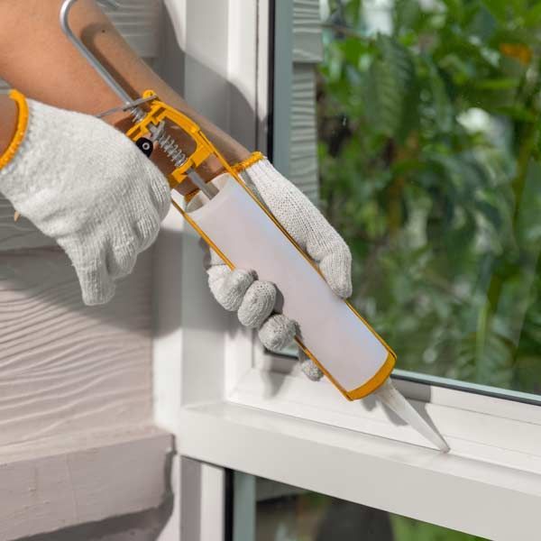 Person caulking a window frame with a caulk gun, wearing gloves, outside, providing Handyman General Maintenance Services