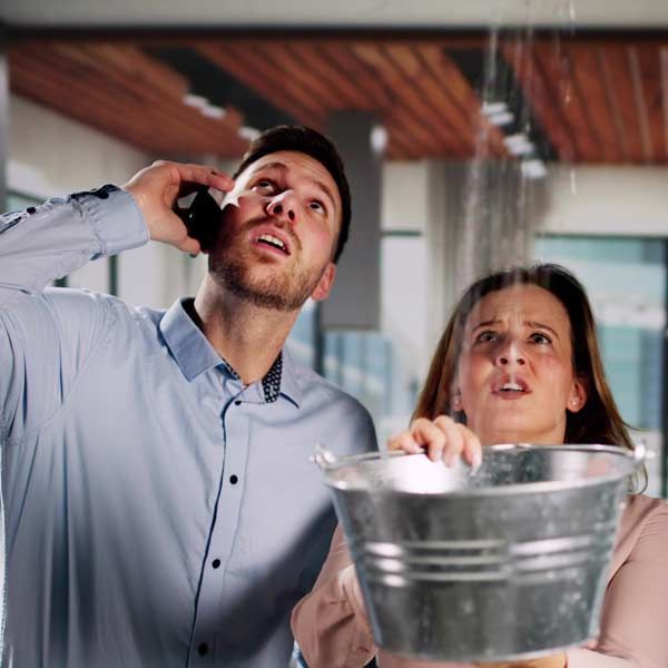 Man on phone, woman holding a bucket, both looking up at a leaking ceiling. Contact Honest Hammer Handyman Repairs and Home Services