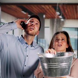 Man on phone, woman holding a bucket, both looking up at a leaking ceiling. Contact Honest Hammer for Home Repairs or Get Started with a Free Home Services Consultation!