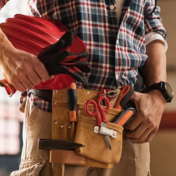 Handyman wearing a plaid shirt and a tool belt, holding a red hard hat and gloves, ready for repair services