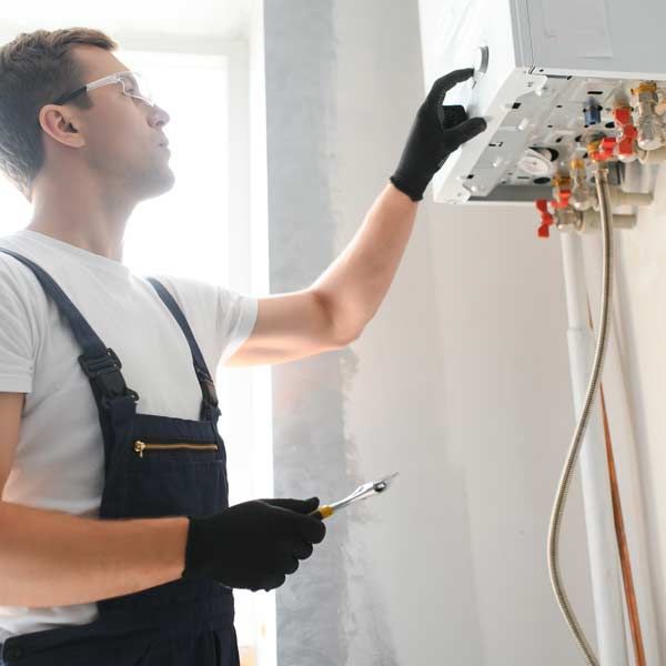 Handyman in blue overalls and gloves, inspecting a white wall-mounted Tankless Hot Water Heater, holding a wrench.