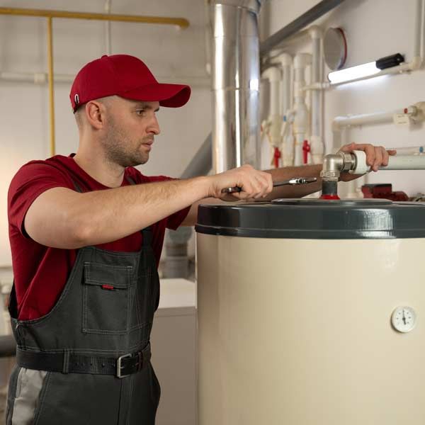 A man in a red shirt and overalls uses a wrench doing hot water heater repairs in a utility room.
