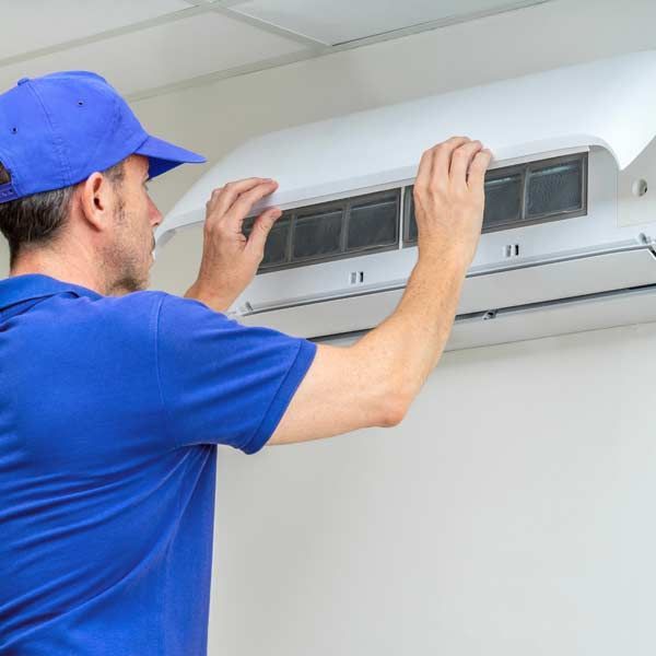 A technician in a blue uniform replacing an air conditioner filter indoors as part of a mechanical updates service