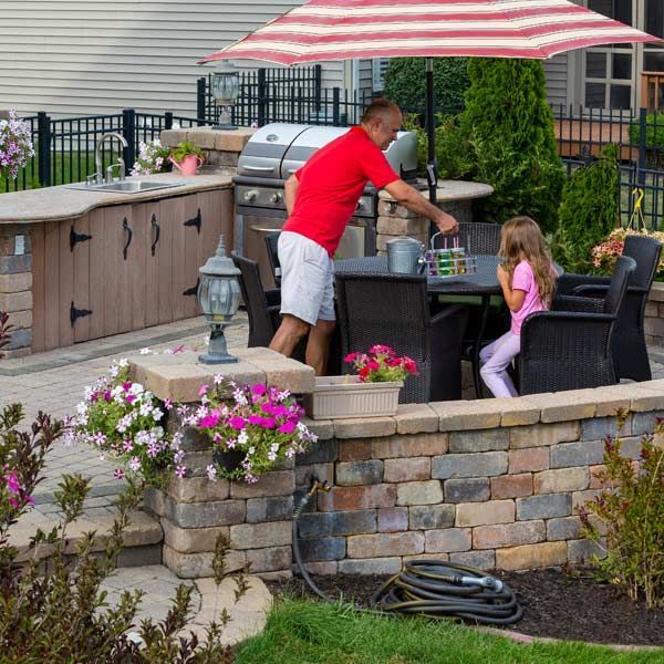 Man and girl at outdoor kitchen with grill, in brick-walled patio with flowers.