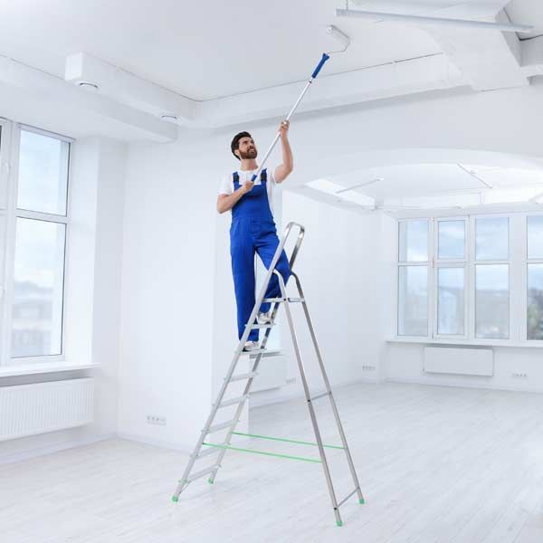Man in blue overalls painting a white ceiling with a roller while standing on a stepladder in a bright room.