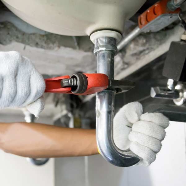 Plumber using a red wrench on a silver pipe under a sink, wearing white gloves.