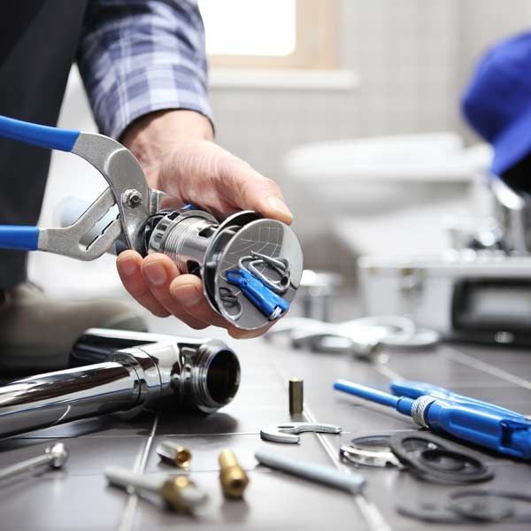 Handyman using pliers to work on sink drain, tools, and parts spread on tile floor Plumbing Repair Services