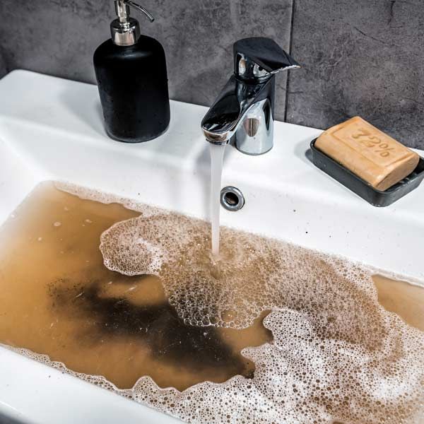 Brown, murky water fills a white sink with a running faucet. Soap and a soap dispenser sit nearby needing Clogged Drain and Toilet Clearing