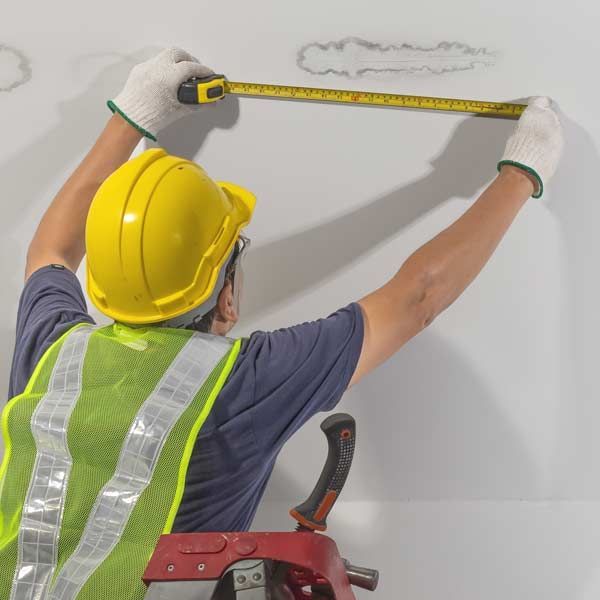 A handyman in a hard hat measures water damage from a plumbing leak on a white wall.