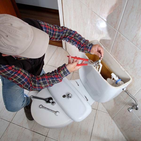 Handyman in plaid shirt repairing a leaking toilet, using pliers. Beige cap, wrenches on the toilet seat.