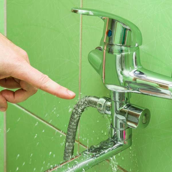 Hand points at a faucet with water leaking from the hose, green tiles background, needing Leaky Faucet and Fixture Repair