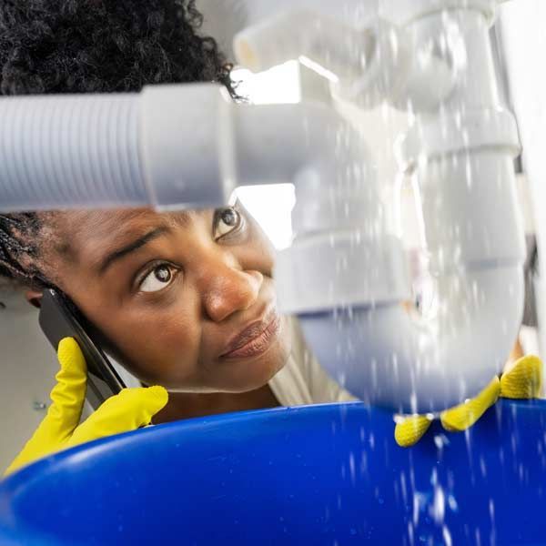 Woman on phone, calling for Leaky Pipe Repairs concerned, watching a leaky pipe over a blue bucket, wearing yellow gloves.