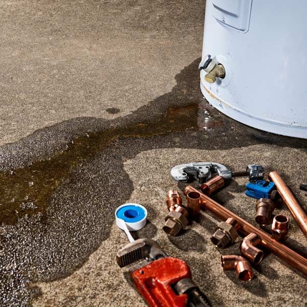 Leaking water heater on concrete floor with handyman's tools, copper pipes, and wrench during Water Heater Repair and Maintenance